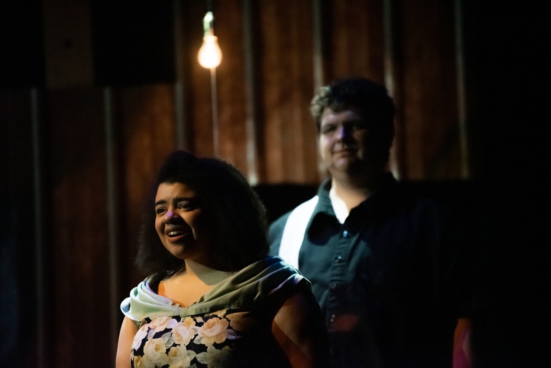  Two performers stand under the illumination of a single light bulb. The performer in the front is viewable from the waist up. She is wearing a flowered dress and green cowl. The figure behind her is wearing a black button up shirt. 