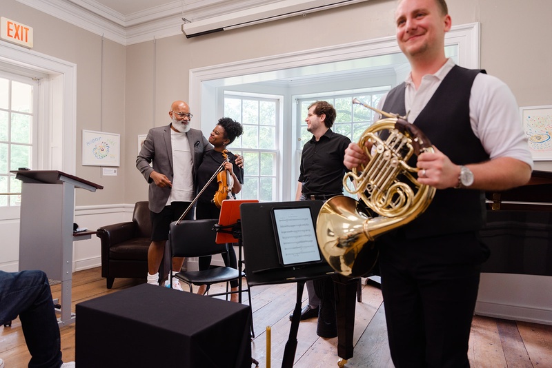  Four performers stand in an all white room with large windows. Two of them are holding instruments, there are a few black chairs and music stands spread out in the room. 