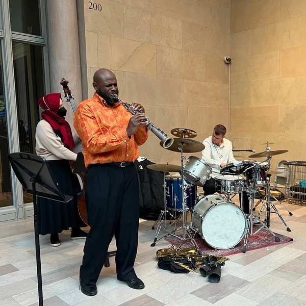  A White man with a beard plays the drums with two other performers. One stands in the front and is playing the saxophone, the other plays the cello. 