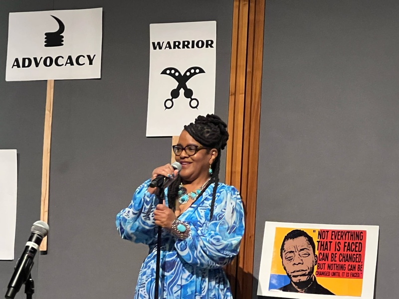  Black woman standing at mic wearing blue dress. There are 3 signs in the background. They read from L-R Legacy, Warrior, image of James Baldwin 