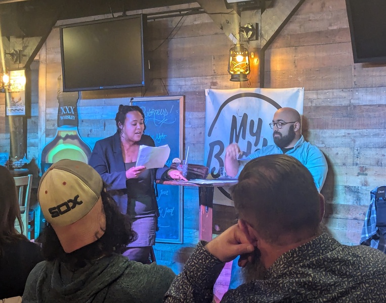  Two performers are standing at a staged area of a bar, reading from a script. One performer is wearing a black dress while the other is wearing a blue button up shirt and glasses. 