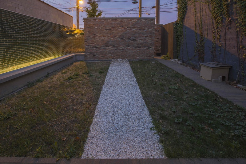  A narrow backyard at dusk features uneven grass on both sides and a central gravel pathway leading to a brick wall. The left wall is adorned with green glass tiles, subtly lit by yellow lights. The right wall has grey tiles with ivy. 