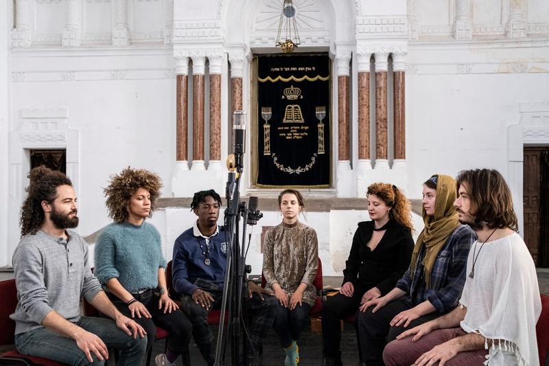  An image of a group of people in a band sitting in a semi circle on a wooden bench surrounding a microphone. Everyone has their hands on their laps as they stare into the distance. 