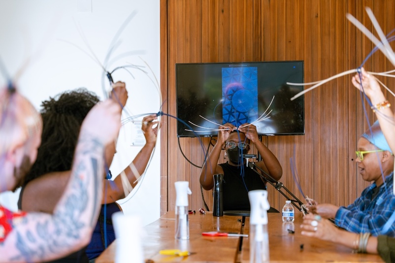  A black woman with glasses, queer presenting with short black hair leads a basket weaving workshop indoors. 