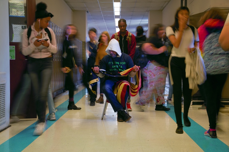  A hooded man, holding an American flag, seated in a high school hallway surrounded by children of multiple genders and races. 