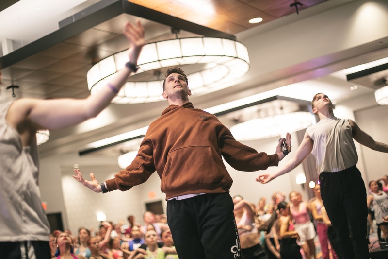  A White/Queer Masc Person with Shorter brown hair posing with their arms outstretched. They are wearing a brown hoodie and black sweat pants. 