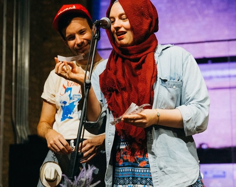  An image of a person speaking into a microphone, reading off a piece of paper. They have light skin and blue/green eyes and are wearing an red hijab 