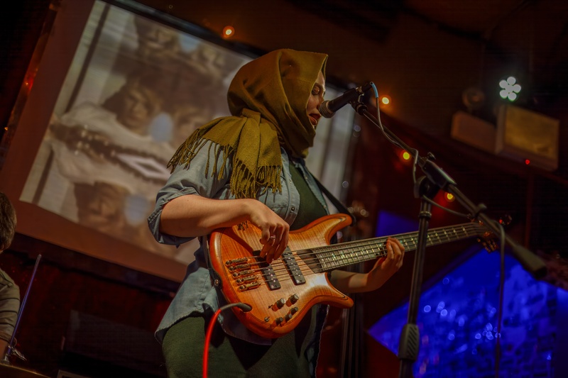 An image of a person playing bass and singing into a microphone. They have light skin and blue/green eyes and are wearing an olive green hijab and blue button down shirt. 
