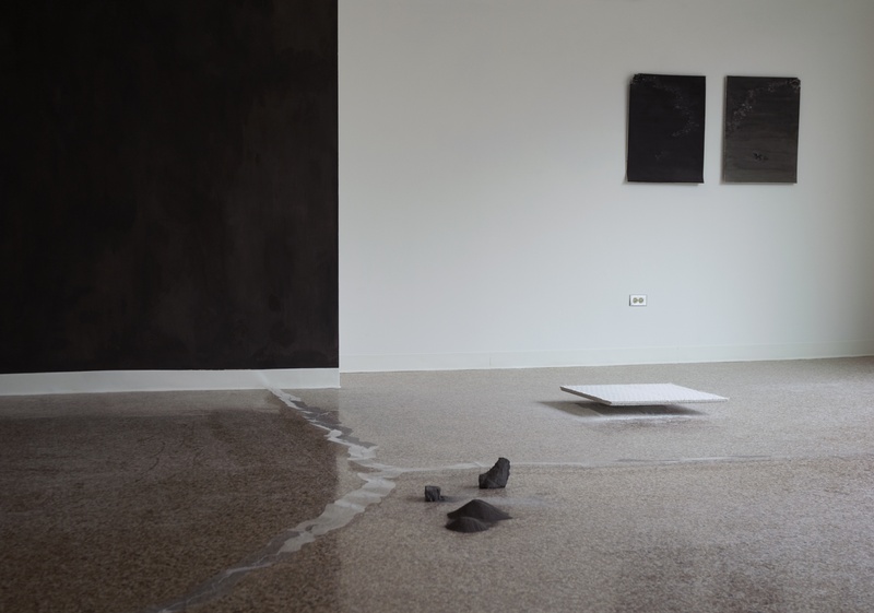  A gallery with a large dark-grey painting on the left wall, flanked by two smaller grey wall works on the right wall. The polished floor has a cracked appearance, with several small grey ash casts and a white ceiling tile platform placed on it. 