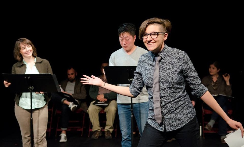  Three performers stand on stage performing a staged reading with music stands. 