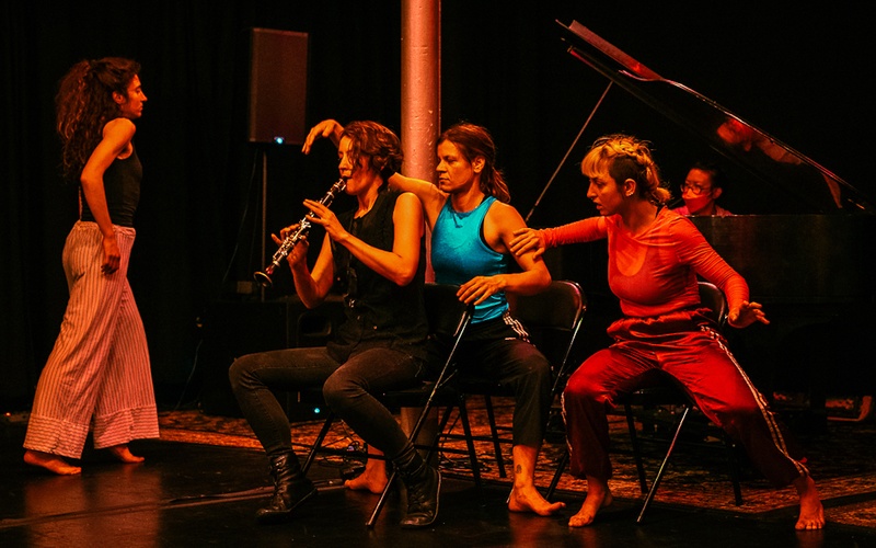  Group of four performance artists on stage, one person plays clarinet, red light hits them all from above 