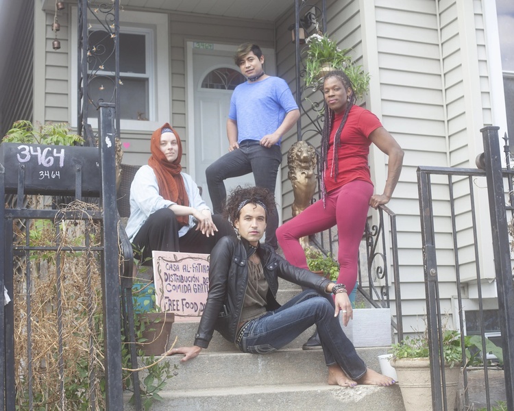  Four people sit and stand on concrete steps outside a house. They are staring a the camera, one individual has They have light skin and blue/green eyes and is wearing a orange hijab.jab 