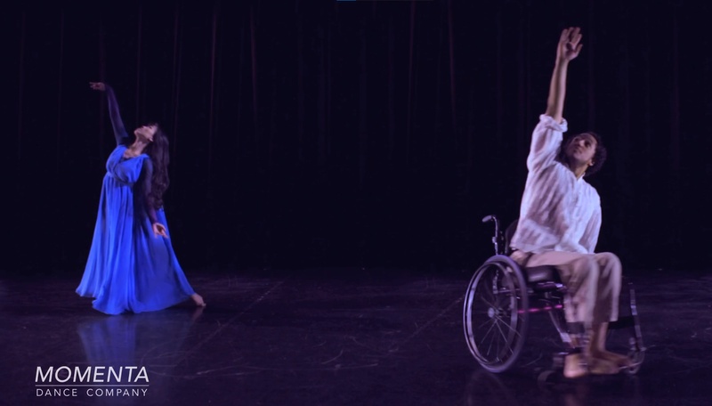  one standing and one dancer in wheelchair dance on dark stage with hands pointing to ceiling 