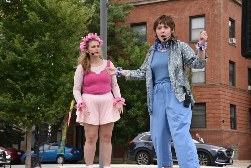  Two performers are standing outside at a street corner. One is wearing a pink dress and flower crown and then other is wearing blue scrubs and a flower necklace. They both have wireless microphones. 