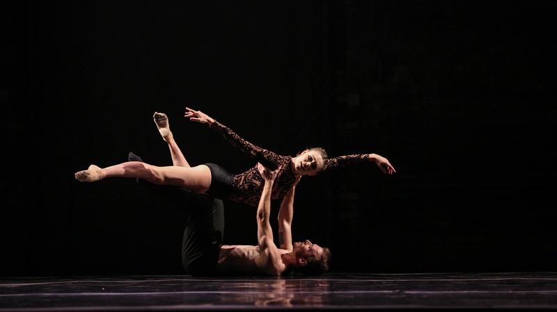  Two performers post mid-dance. One is on the ground lifting the other from their arm pits. The performer in the air stretches their arms out. They are both wearing black. 