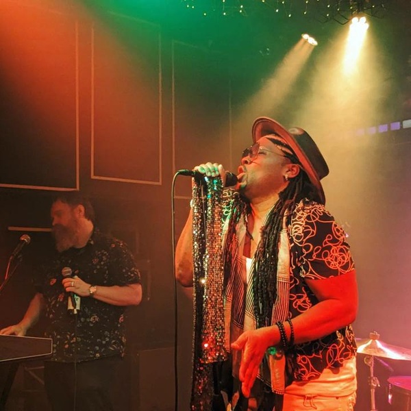  A Black/queer performer with Dreadlocks stands at a microphone, singing. They are wearing a brimmed hat and are holding a silver sequined cloth over the microphone. 