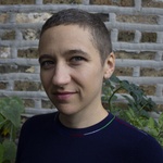 A light-skinned person with very short hair and a neutral expression stands in front of a brick wall with plants in the background. She wears a dark blue sweater with a crew neck collar..