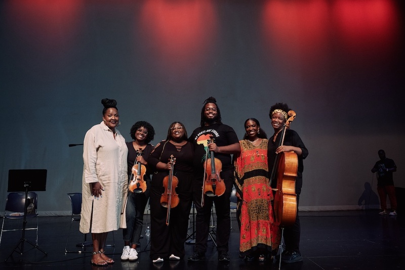  Six people stand on a stage. They are holding musical instruments, three violins and one cello. They are all smiling at the camera, most are wearing black but one person is in all white. 