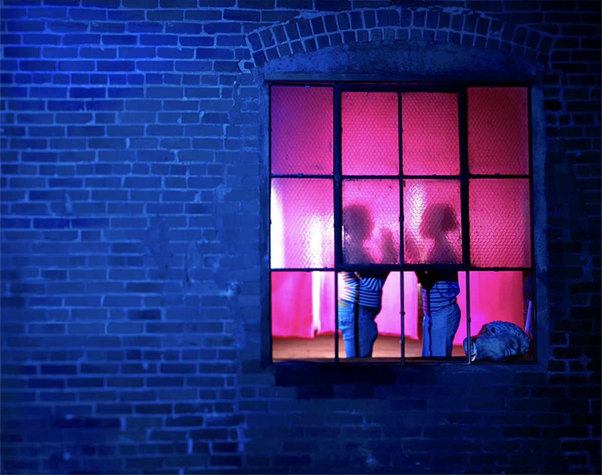 Image saturated in blue excepted for an interior window which appears deeply pink. Inside the window are two profiles of people playing a hand-clap game.